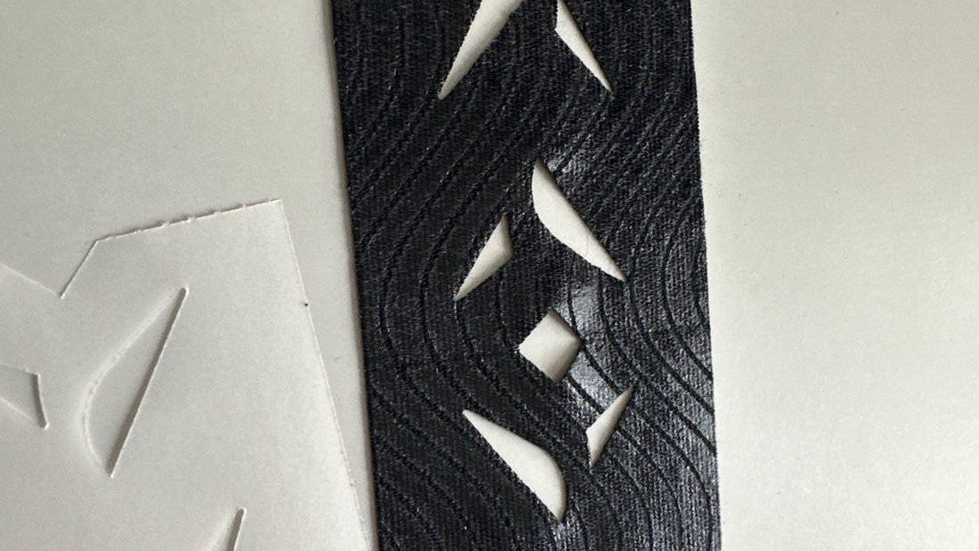 Close-up of a black pre-cut kinesiology tape strip with diamond cut-outs, adhesive side up beside its backing paper